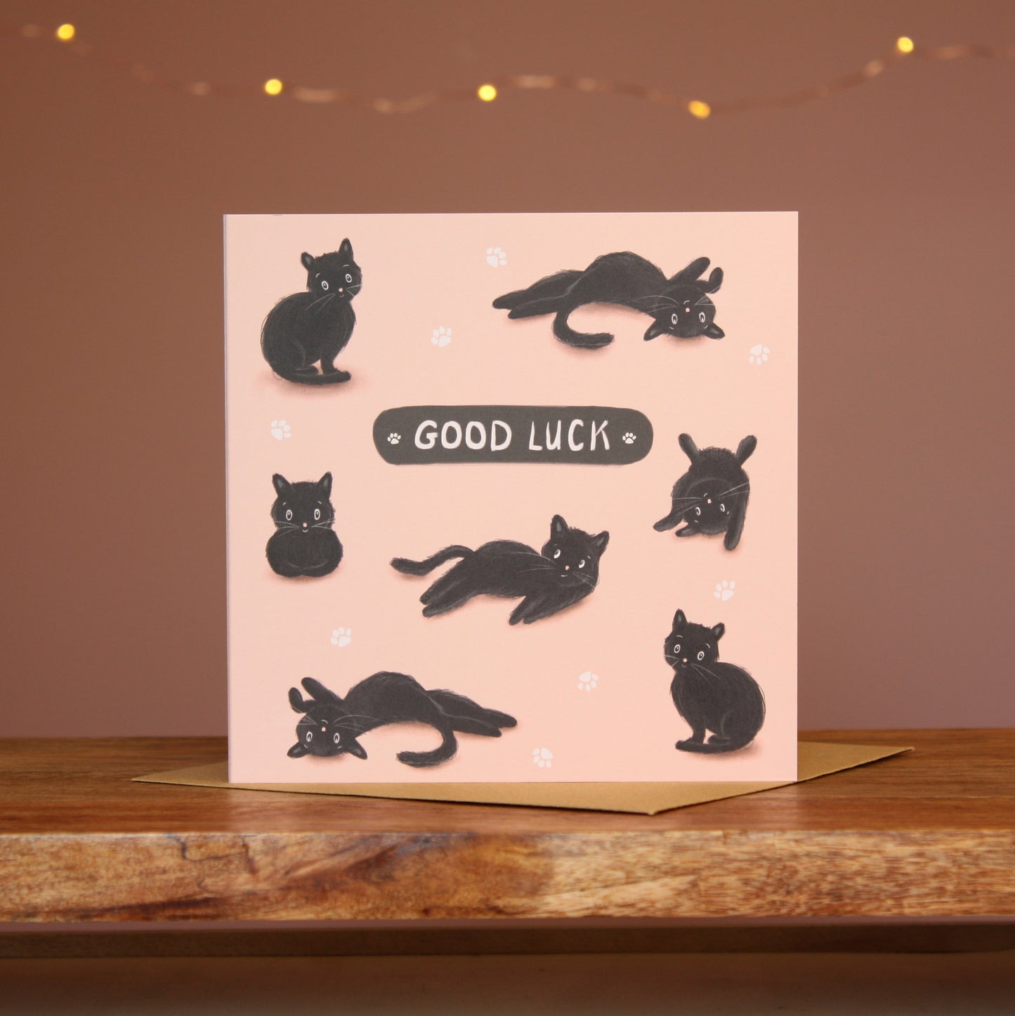 A square card featuring an illustration of a black cat in various 'cat' positions against a pale pink background with the words'Good Luck'.