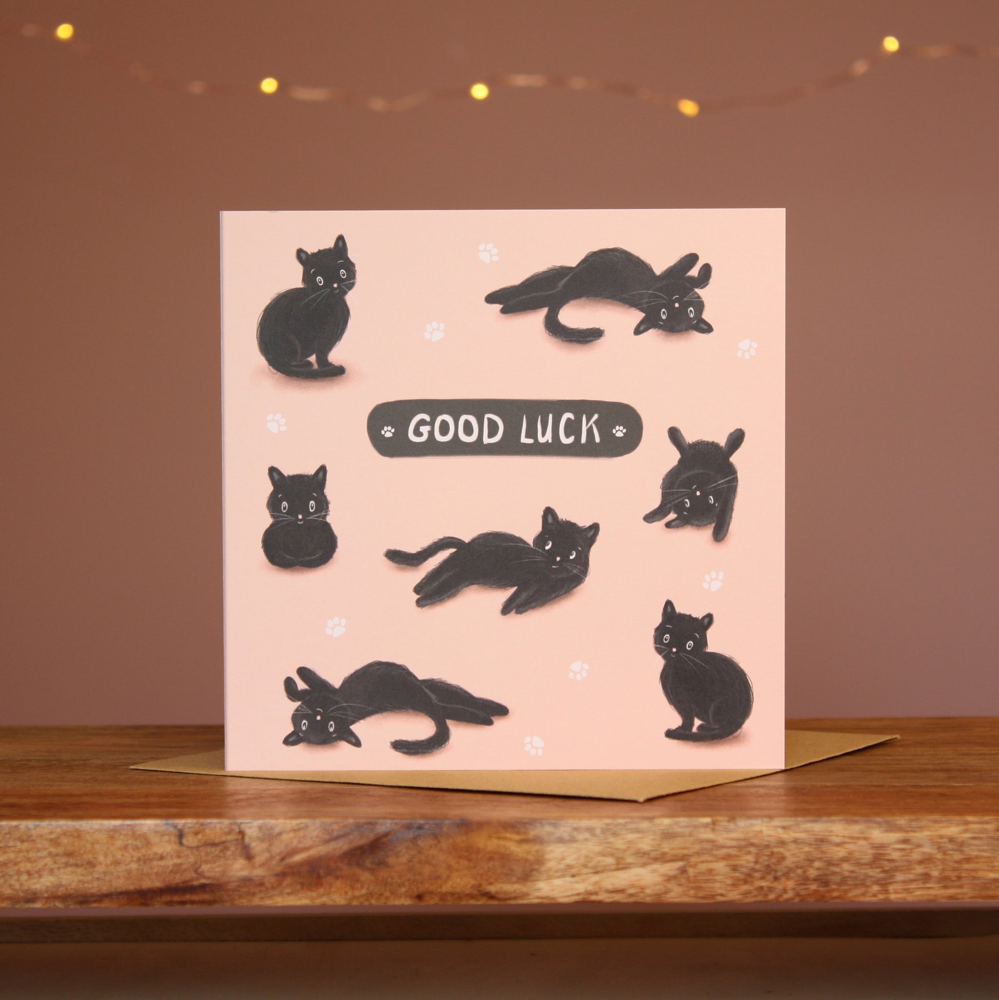 A square card featuring an illustration of a black cat in various 'cat' positions against a pale pink background with the words'Good Luck'.