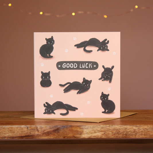 A square card featuring an illustration of a black cat in various 'cat' positions against a pale pink background with the words'Good Luck'.