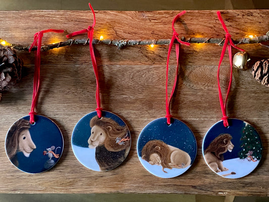 A set of four round ceramic Christmas decorations with red velvet ribbon. Ornament 1 shows a mouse giving a lion a gift, ornament 2 is of a lion and a mouse in a snow blizzard, ornament 3 is of a lion and mouse sleeping in the snow and ornament 4 is a lion watching the mouse decorate the Christmas tree.