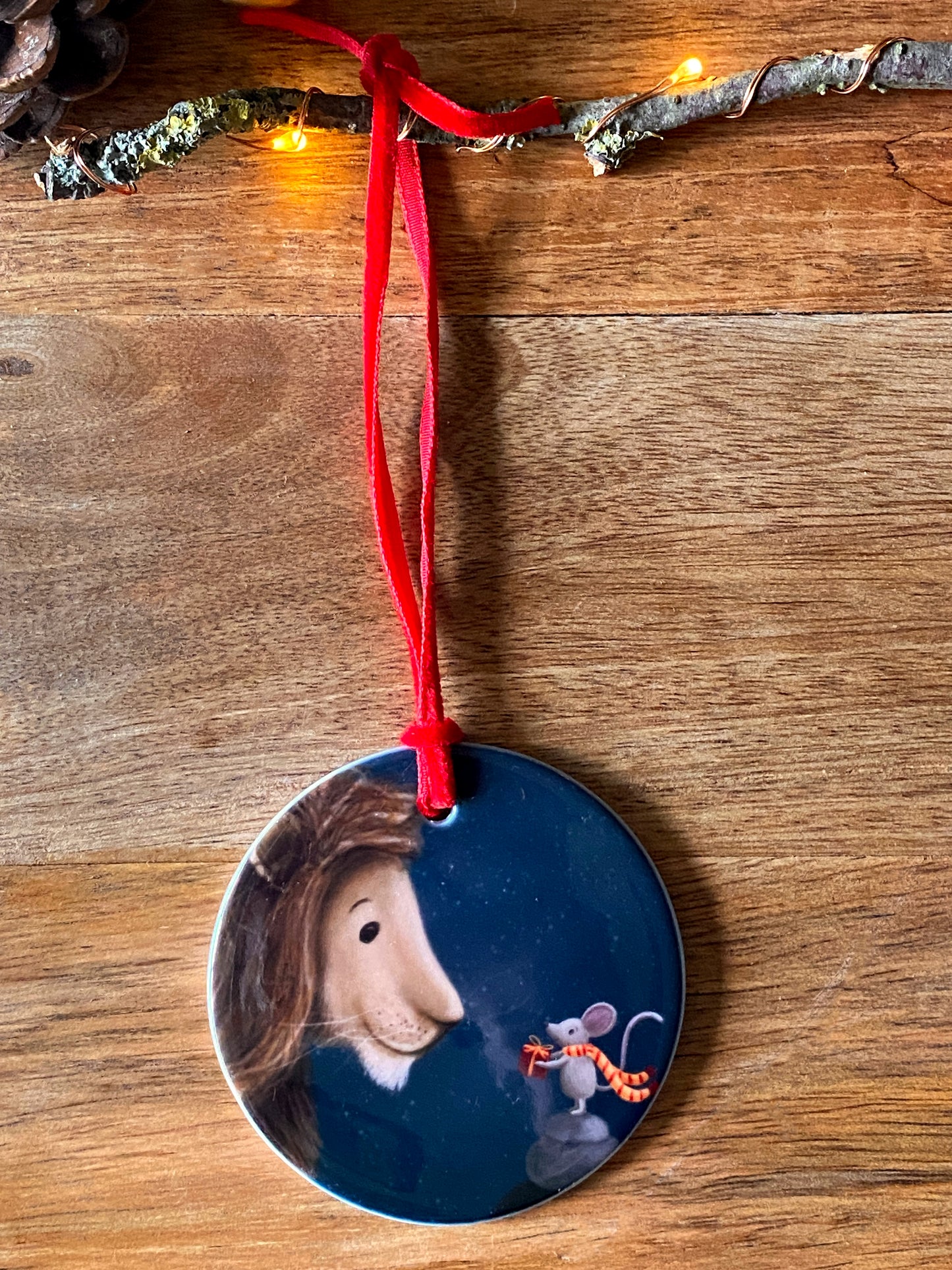 Round ceramic Christmas tree decoration with red velvet ribbon showing an illustration of a mouse giving a lion a present against a night lit sky. 