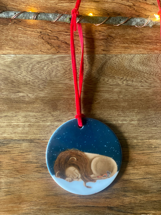 Round ceramic Christmas tree decoration with red velvet ribbon showing an illustration of a lion and mouse asleep in the snow against a snowy night lit sky. 