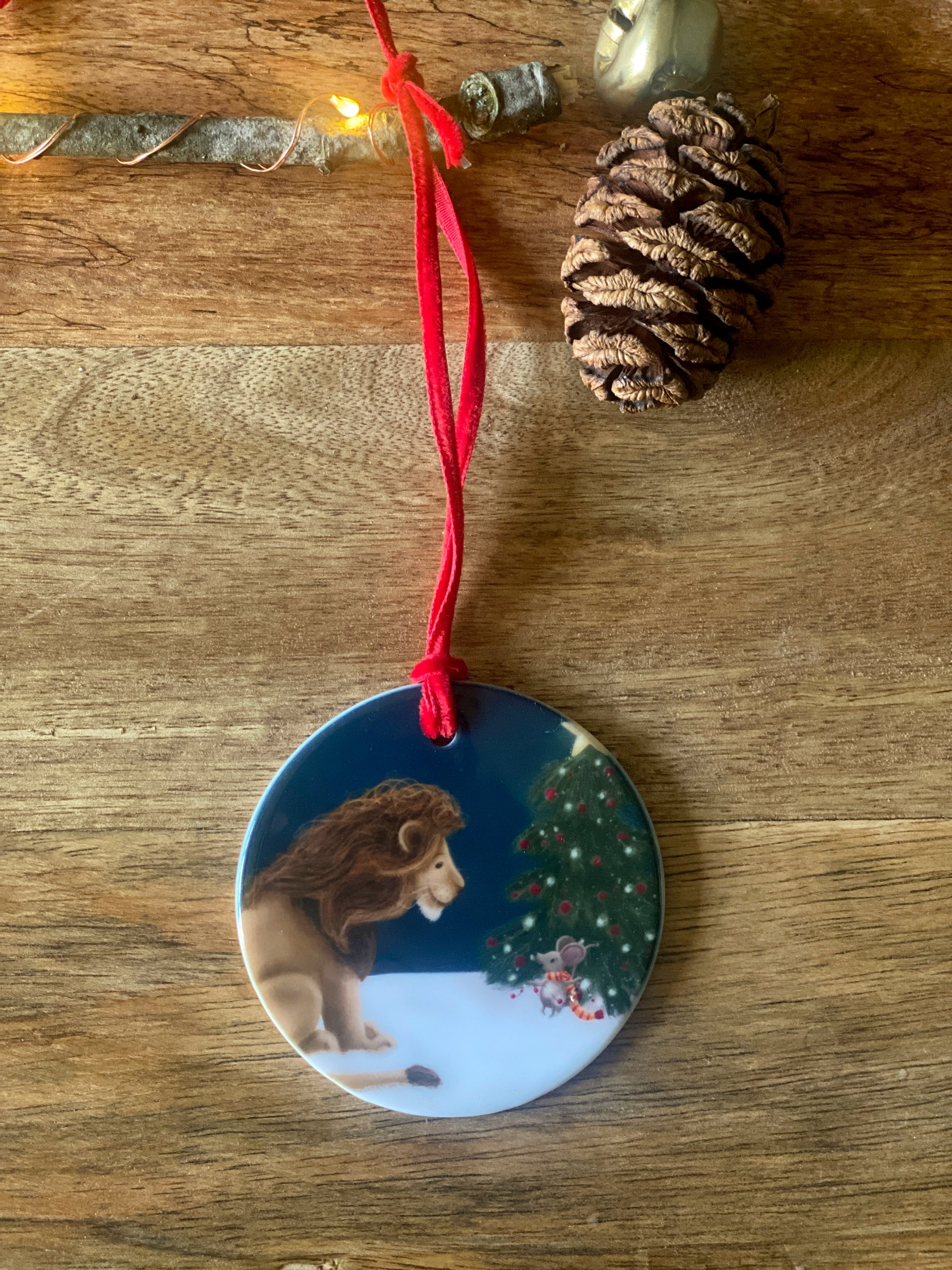 Round ceramic Christmas tree decoration with red velvet ribbon showing an illustration of a lion watching a mouse decoration the Christmas tree against a night lit sky. 