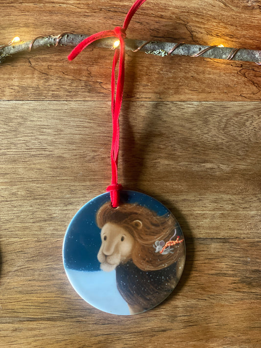 Round ceramic Christmas tree decoration with red velvet ribbon showing an illustration of a lion and mouse walking in a snow blizzard against a night lit sky. 