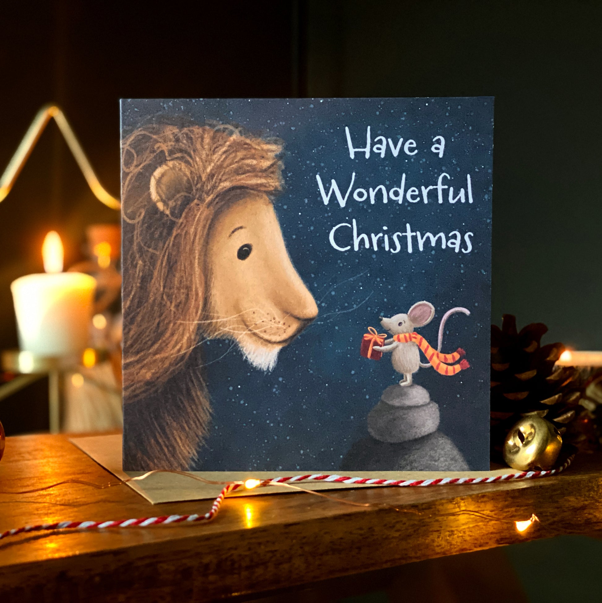 A square Christmas card showing an illustration of a mouse giving a lion a present against a night lit sky. 