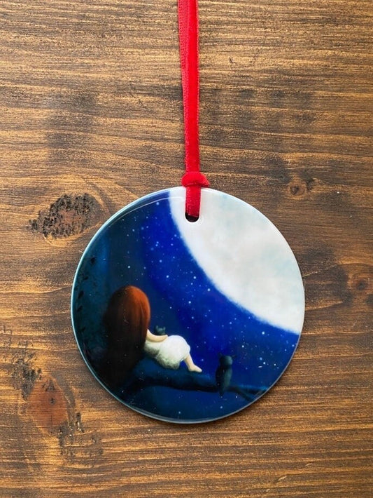 A round flat ceramic ornament, hanging from a red velvet ribbon, with an illustration of a girl sat in a tree with a cat and owl looking at the moon in a star lit sky. 